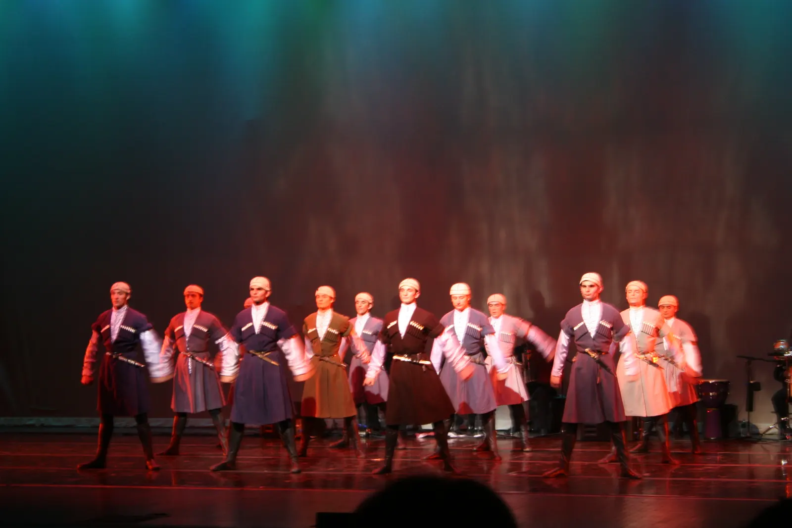 A row of Georgian male dancers in long coats and caps on a stage under red and teal light