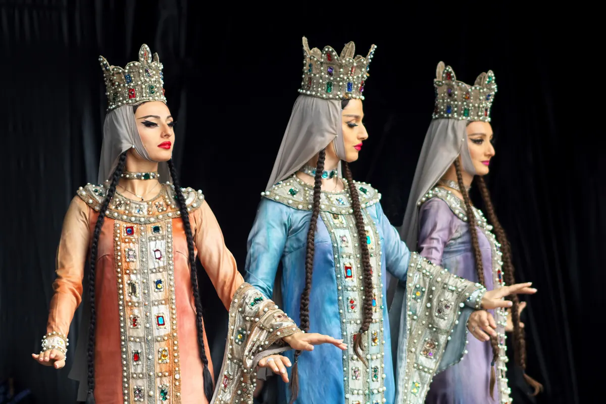 Three Georgian women dancers in profile, in jewelled crowns, veils and long beaded sleeves, on a dark stage