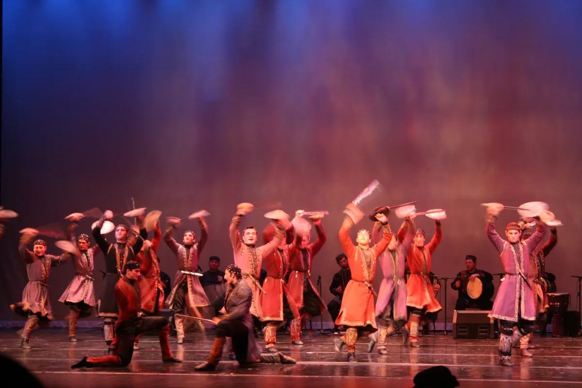 Georgian male dancers in red and purple coats with arms raised, mid-spin, on a stage lit in blue and red