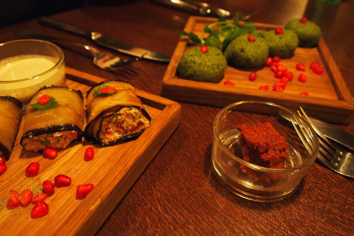 Eggplant rolls and green pkhali balls with pomegranate seeds on a wooden board, beside a small jar of red ajika