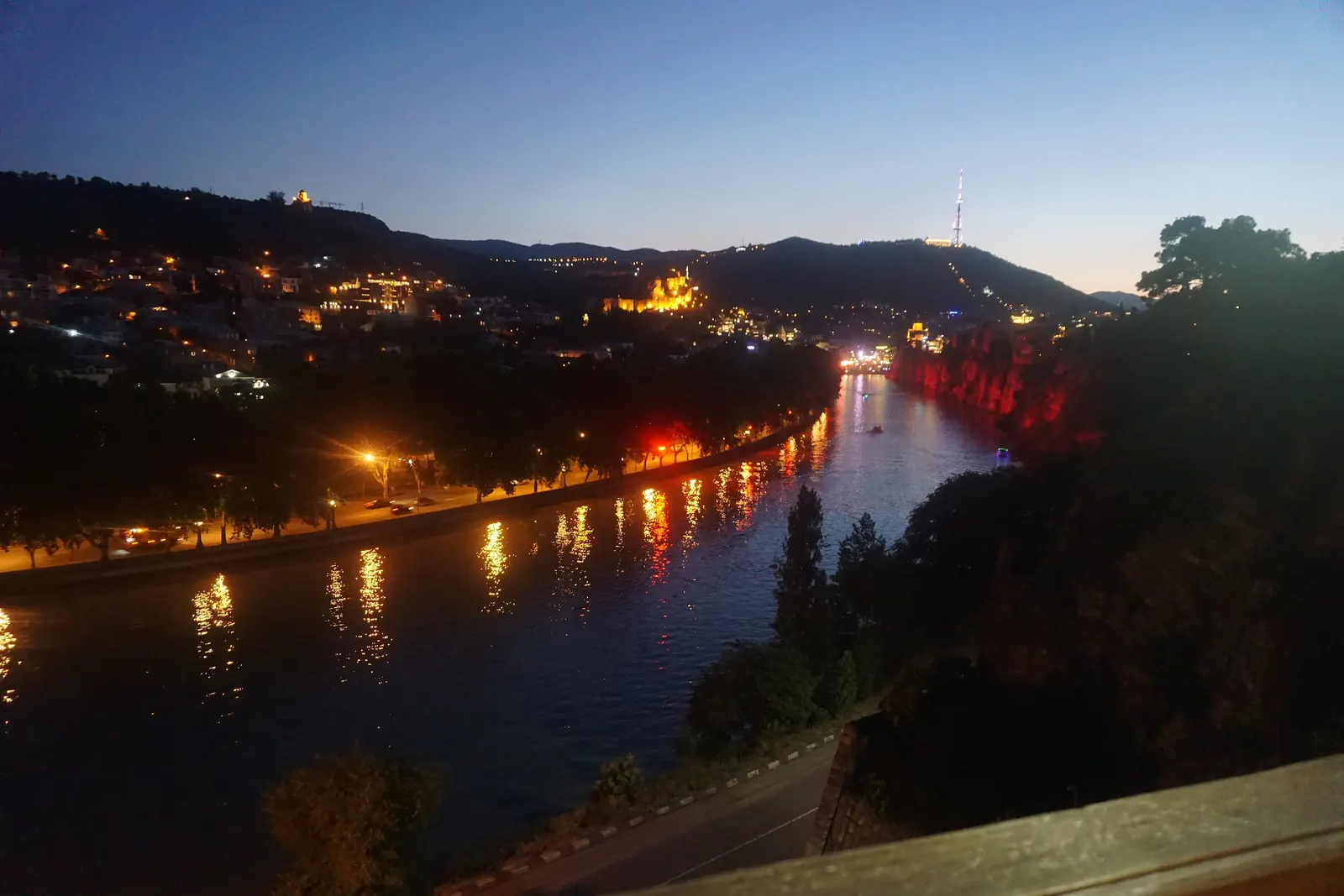 The Kura river in Tbilisi at dusk, with lit-up hillsides and the city's lights reflected in the water