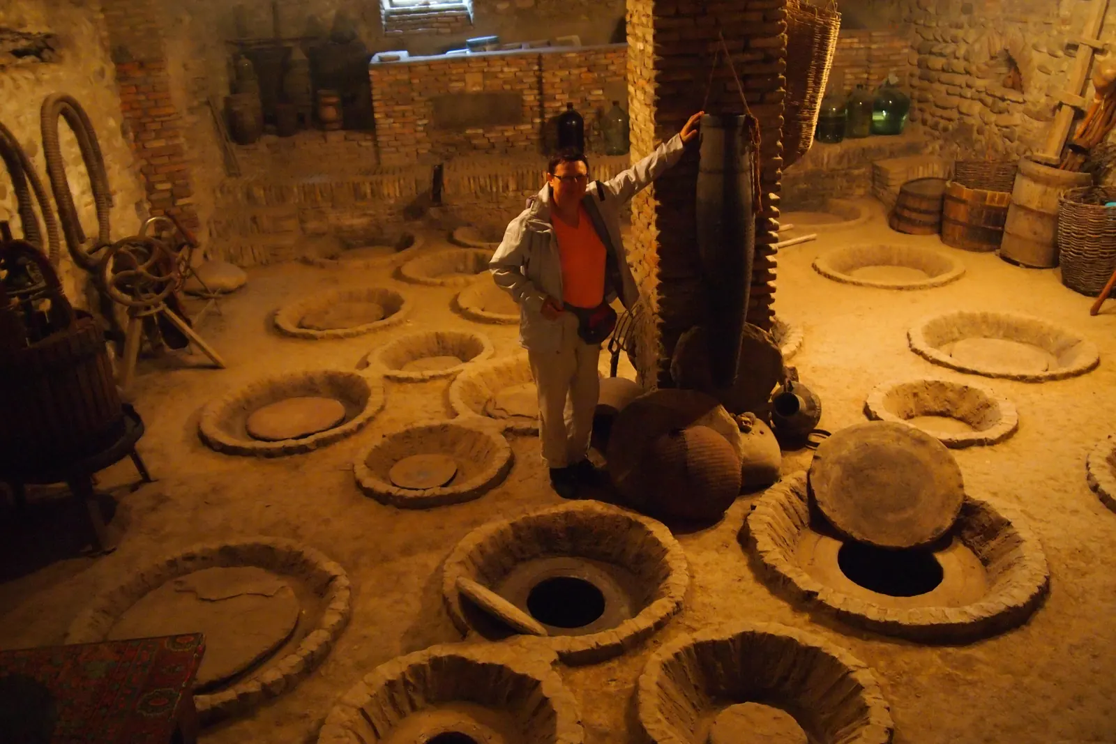 Wide clay rims of qvevri jars set level into the stone floor of a Georgian winery cellar, with barrels and tools behind