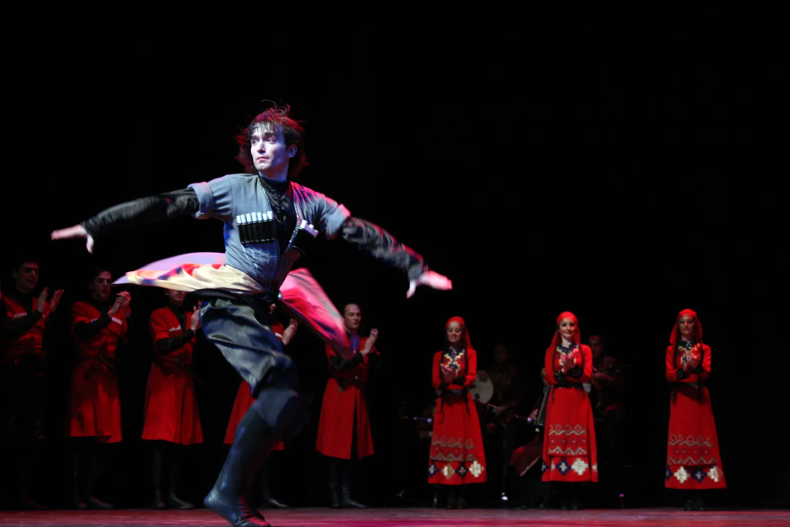 A male dancer in a dark Georgian coat leaping across a stage while women in red dresses stand in a line behind him